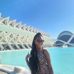 A young black woman in front of the Museum of Arts and Science in Valencia. She is wearing a floral dress, dark sunglasses and has long black braids.