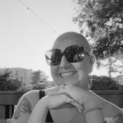 image of a white feminine person with buzzed hair, big sunglasses, smiling outside.