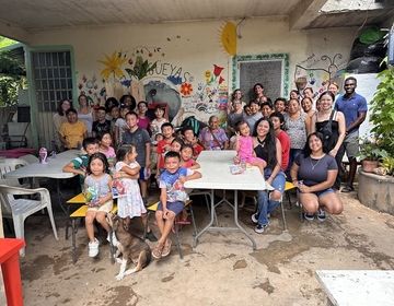Group of children, teens and mothers stand and sit around tables, posing for a group photo. 