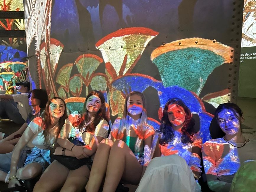 A group fo HSSA Language and Culture students sit on the floor against the wall to enjoy the projections. A colorful pattern of organic shapes is projected on the wall behind them. 