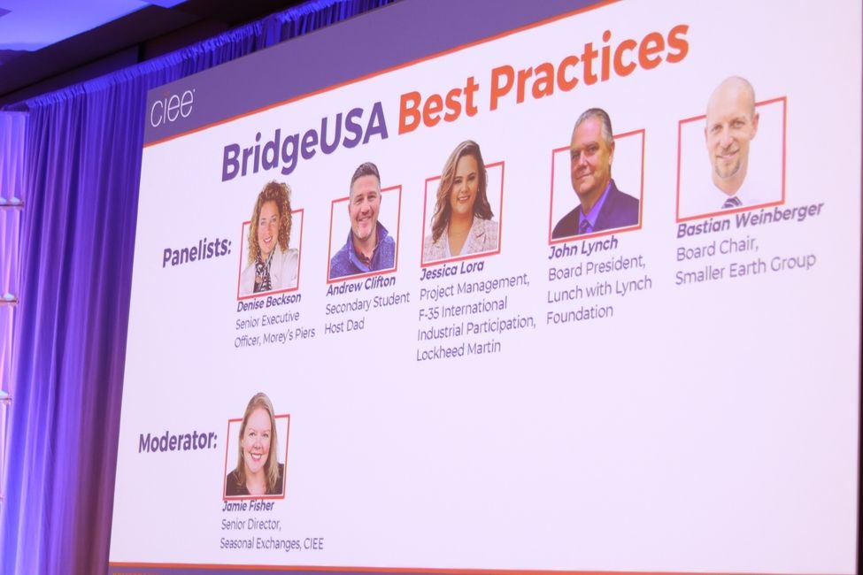 A shot of a large screen that shows a line up of panelists and a header that says BridgeUSA Best Practices