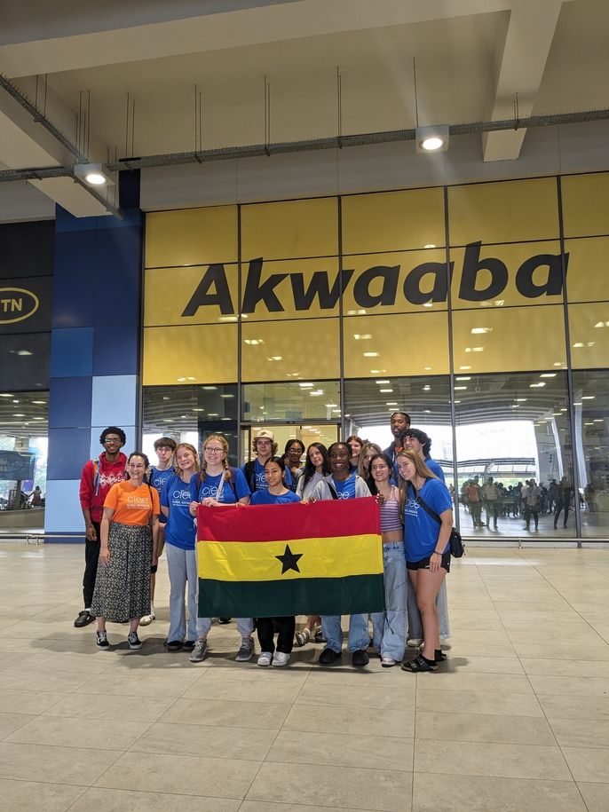 Participants of the 2024 High School Summer Abroad program take a photo in front of the Akwaaba sign at the Kotoka International Airport. A few of them standing in the front row hold up the Ghana national flag. 