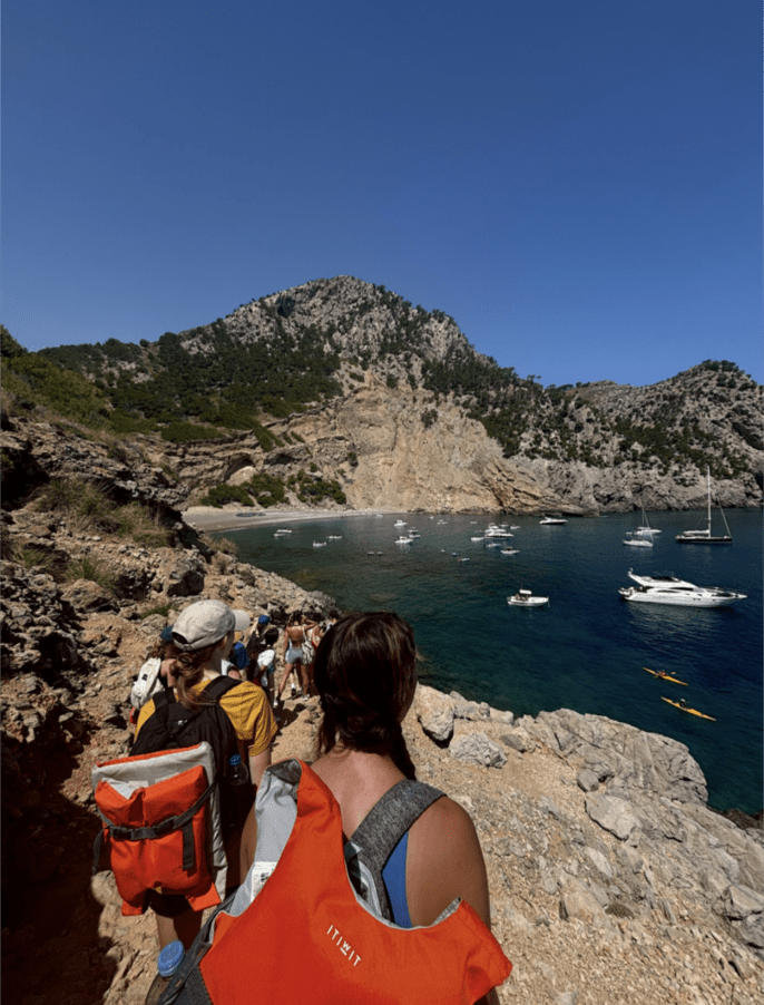 People with backpacks on walk along a rocky trail with a hidden cove in the background and rocky cliffs on the other side of the cove