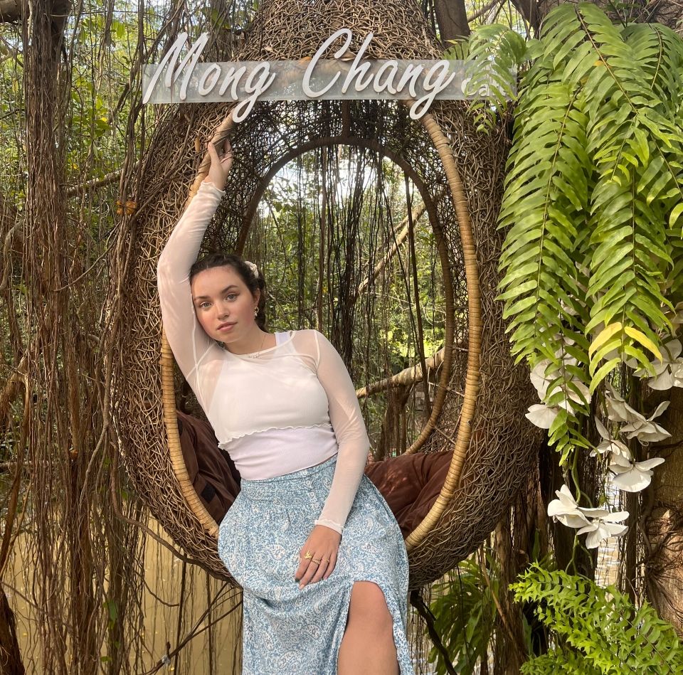 Young woman in a white shirt and blue skirt poses in a hanging chair that reads "Mong Chang"