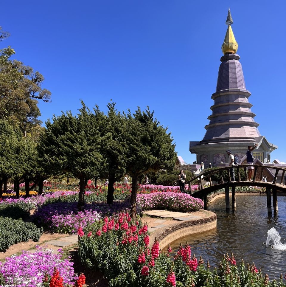 Purple pagoda in the background of a lush garden with purple and red flowers and a wooden bridge over a small creek