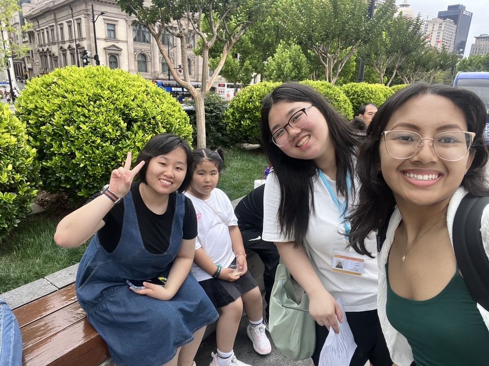 Two CIEE high school students smile next to a mother holding up a peace sign with a child sitting by her side. Beyond a row of bushes are historic buildings of the Bund. 
